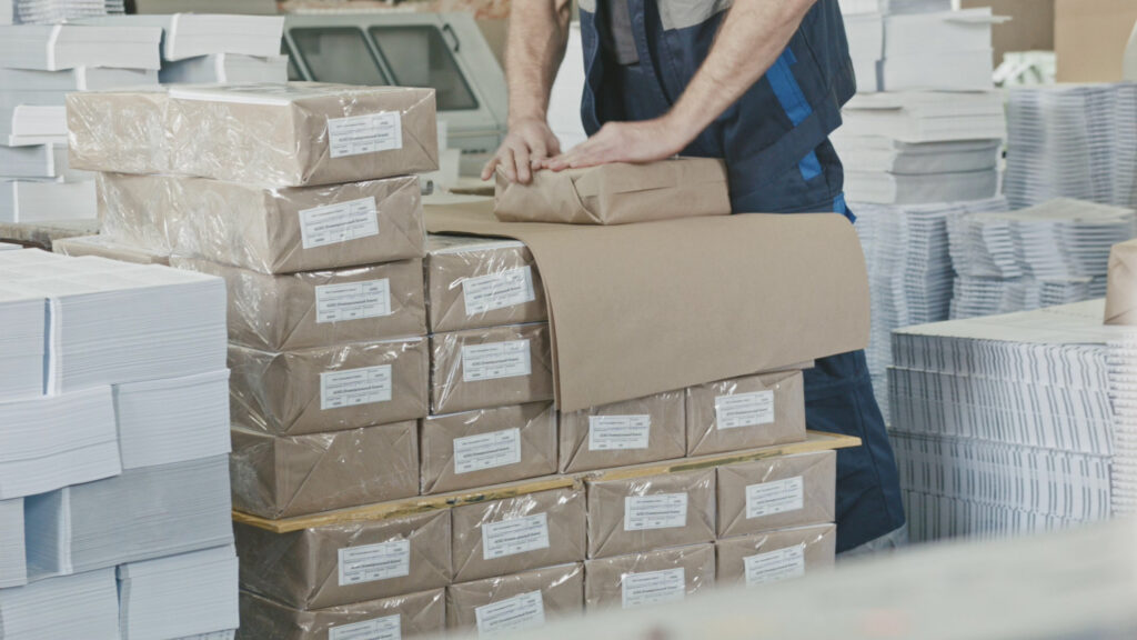 A factory worker is wrapping a package in brown paper on top of a stack of neatly packed and shrink-wrapped bundles of paper. The surrounding area is filled with stacks of printed sheets and paper reams, indicating a print shop or paper processing facility. The worker is dressed in a blue uniform, focused on preparing materials for delivery or shipment.