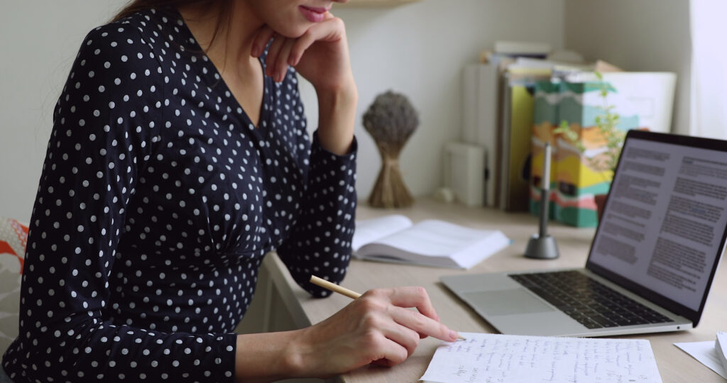 A woman in a black polka dot blouse sits at a desk, thoughtfully writing notes in a notebook with a pencil, figuring cost to serve. An open laptop displaying text is in front of her, and open books, documents, and a small plant are visible in the background, creating a focused and studious work environment.