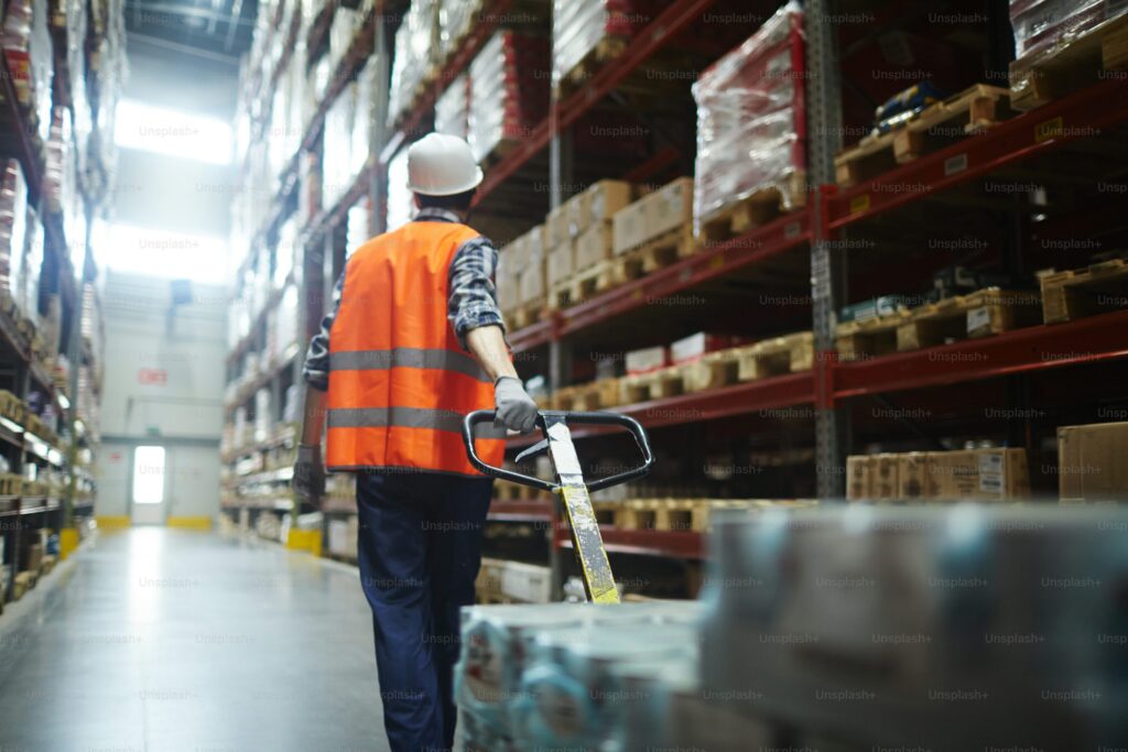 Warehouse worker in an orange safety vest and hard hat pulling a loaded pallet jack through tall warehouse aisles filled with stocked shelves. The image emphasizes order picking efficiency and workflow speed—key considerations in calculating how many orders a team should pick per hour.
