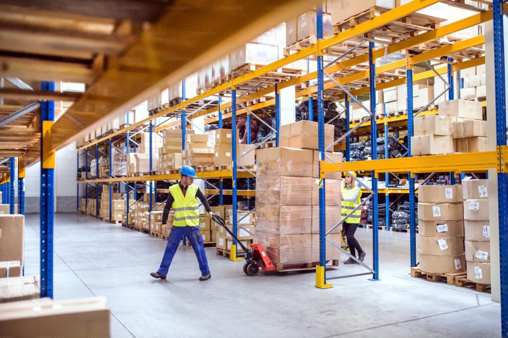 Two warehouse workers in high-visibility vests and hard hats moving a large pallet of boxes with a manual pallet jack through wide warehouse aisles. The scene illustrates order picking productivity and workflow efficiency—important factors in determining how many orders a team should pick per hour.