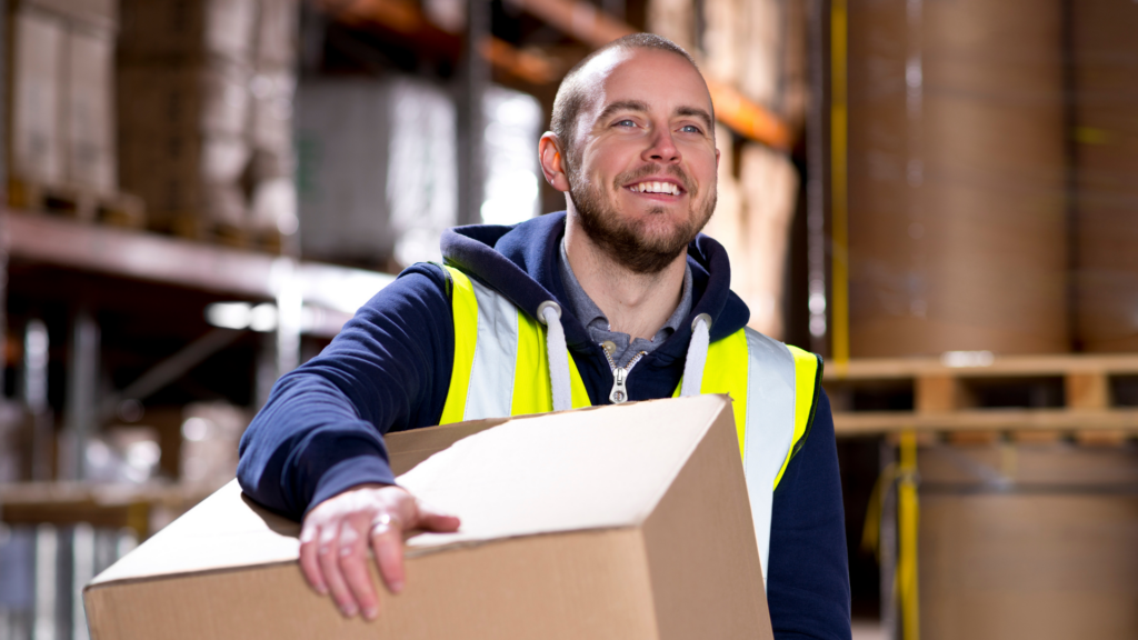 Warehouse associate wearing a high-visibility vest carrying a large box through a warehouse aisle.