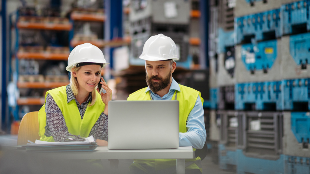 Two warehouse supervisors in safety vests and hard hats reviewing operations data together on a laptop.