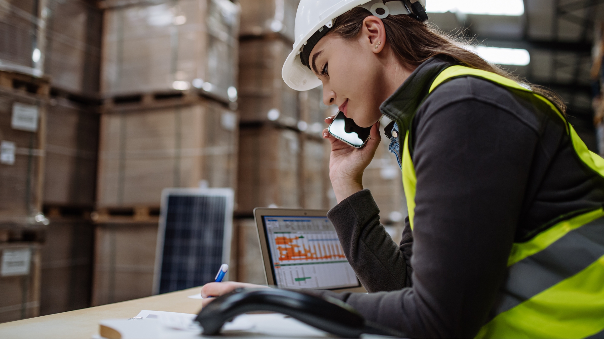 Warehouse operations manager in a hard hat and hi-vis vest reviewing labor data on a laptop while on a phone call on the warehouse floor