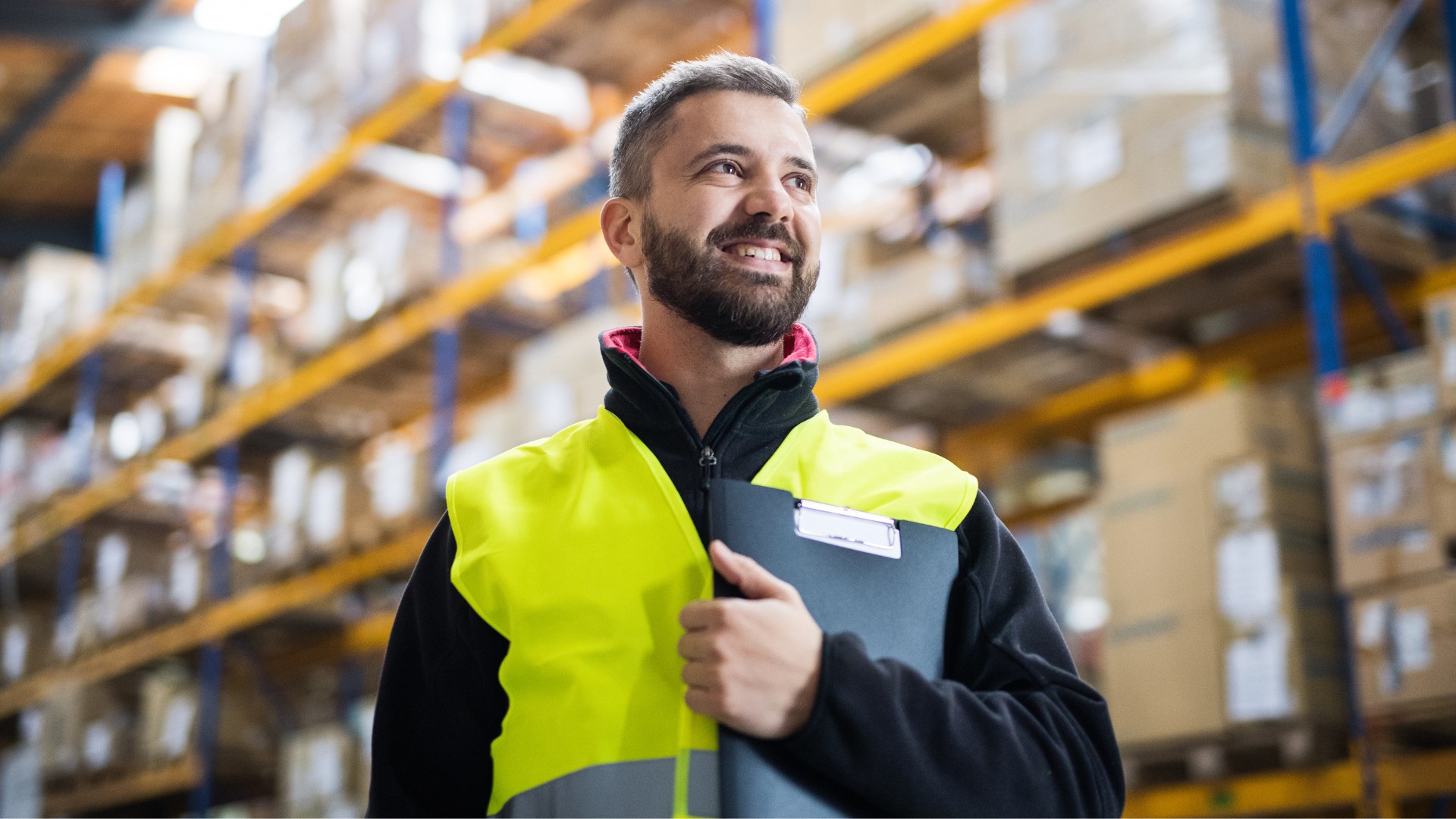 Warehouse supervisor in a high-visibility vest holding a clipboard and surveying the distribution center floor