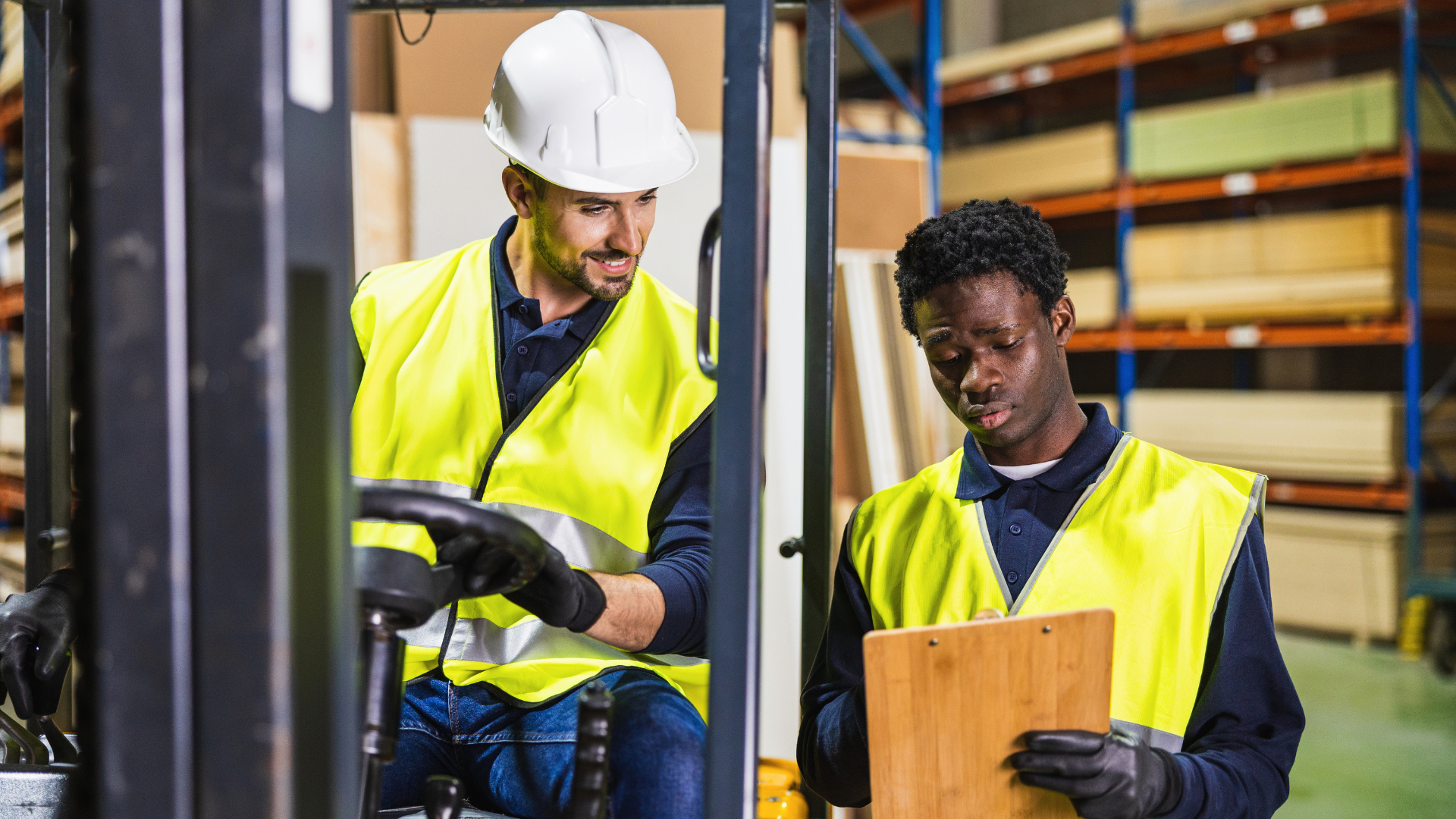 Warehouse supervisor on a forklift reviewing tasks with a coworker holding a clipboard in a distribution center